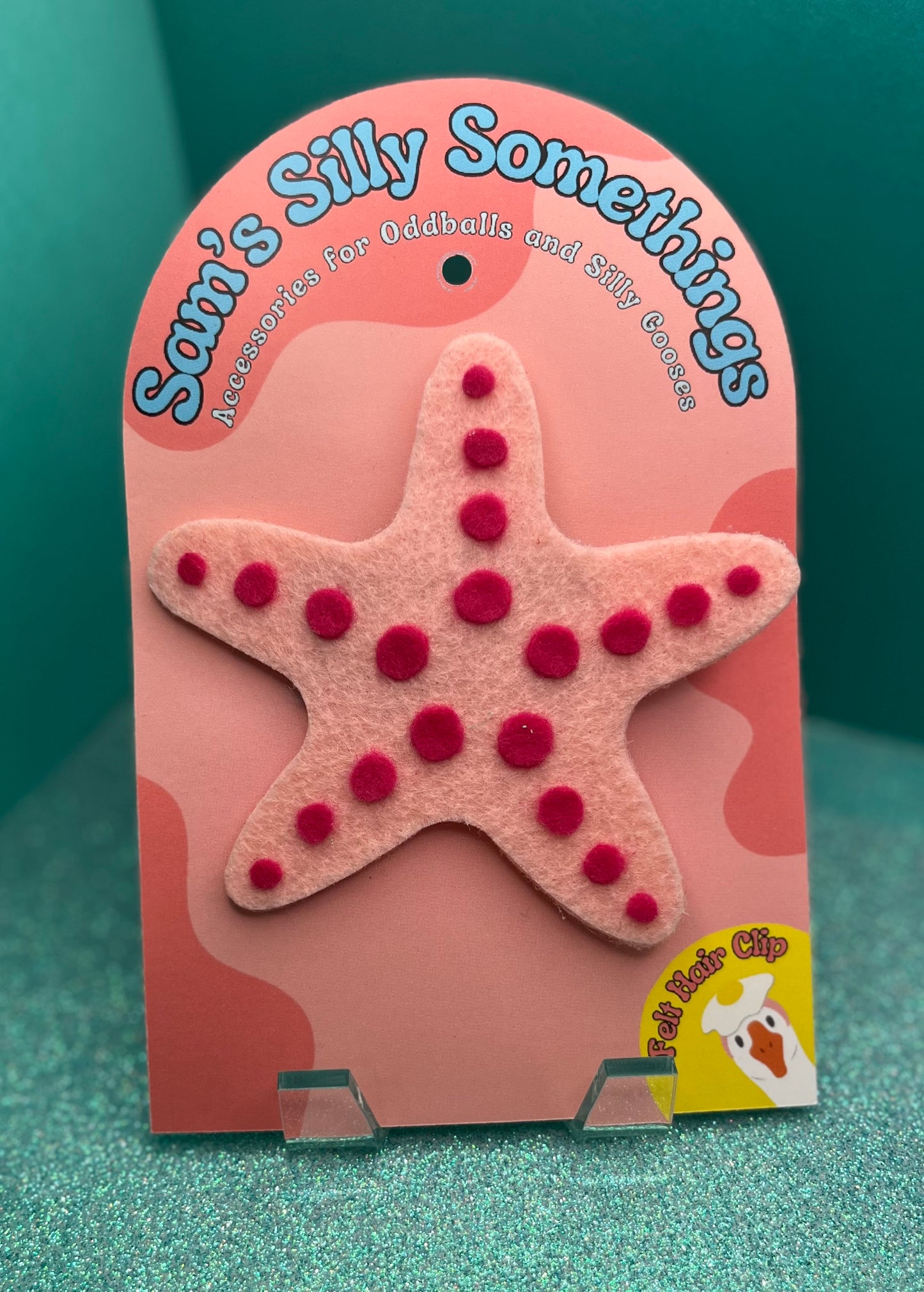 Starfish-shaped hair clip with red pom-poms on a pink card with 'Sami's Silly Somethings' branding.