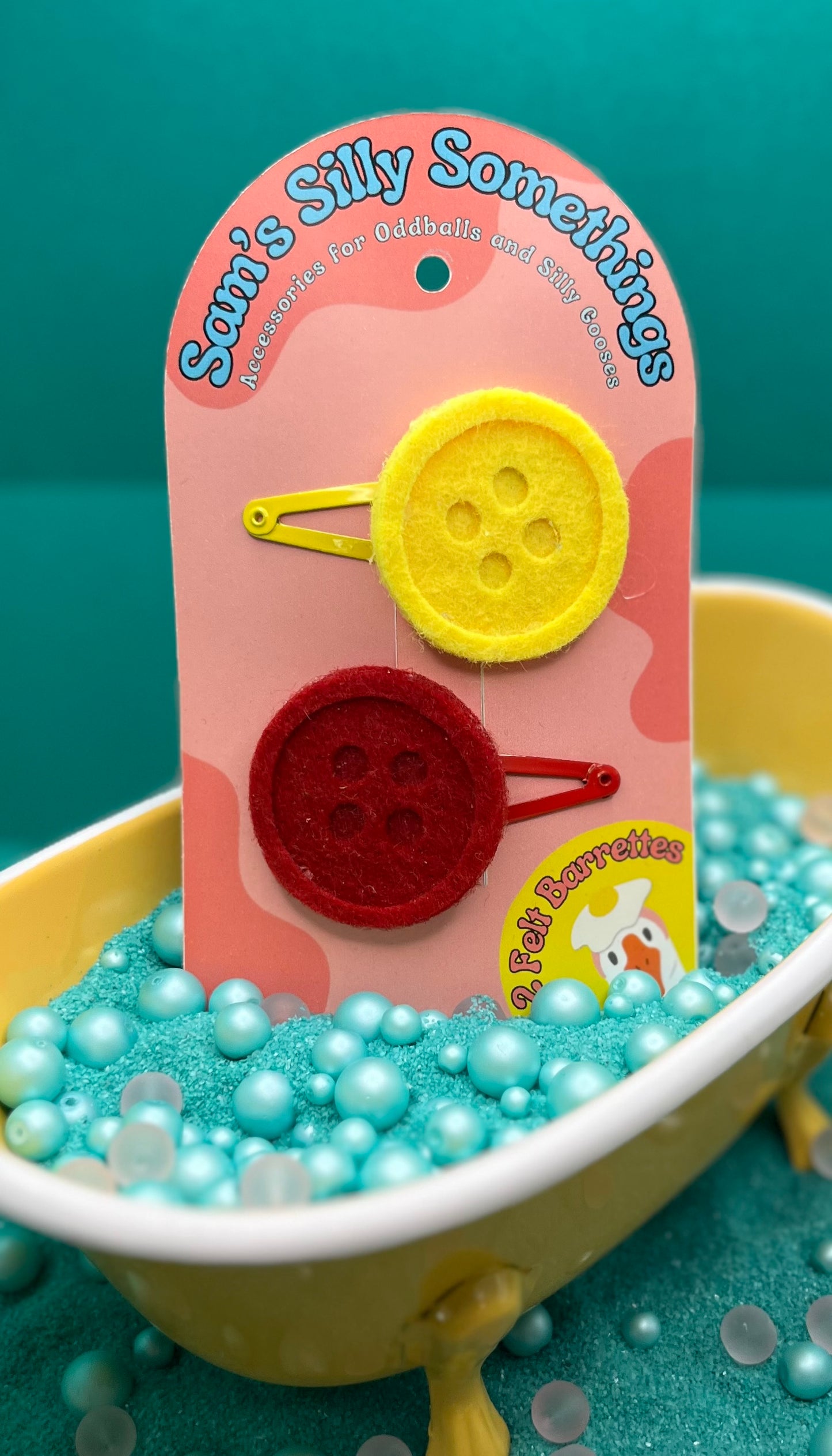 Two button barrettes, one red and one yellow, attached to a nickel plated metal clip, displayed on a packaging card with a playful design.