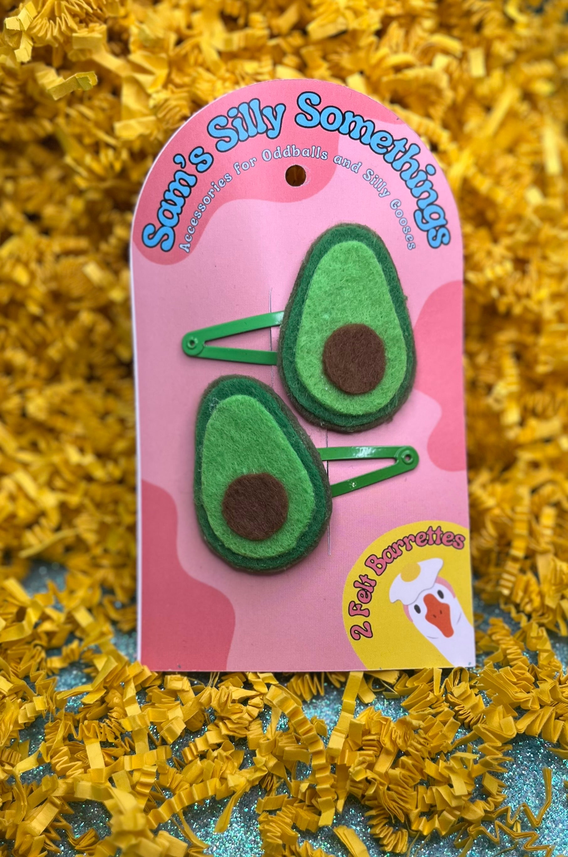 Two green avocado-shaped hair barrettes attached to a pink cardboard backing with the text 'Sam's Silly Somethings'. The barrettes are designed to look like avocados with a brown pit in the center.