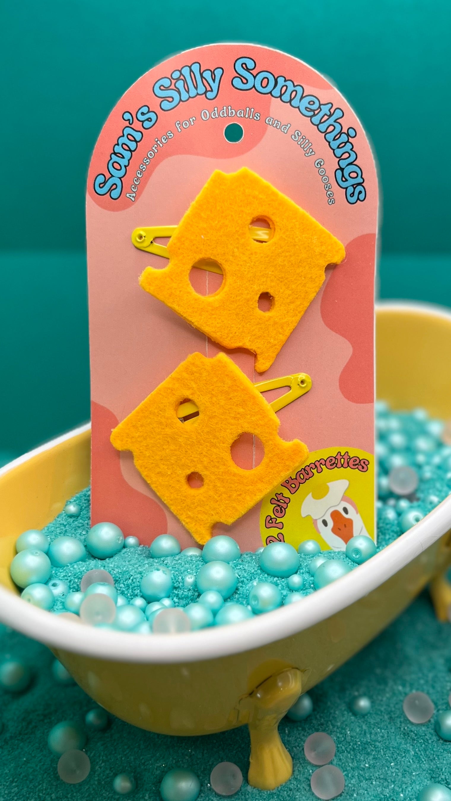 Two yellow cheese-shaped barrettes attached to a pink backing card, presented in a yellow bathtub filled with blue beads, with a 'Sam's Silly Somethings' label in the background.