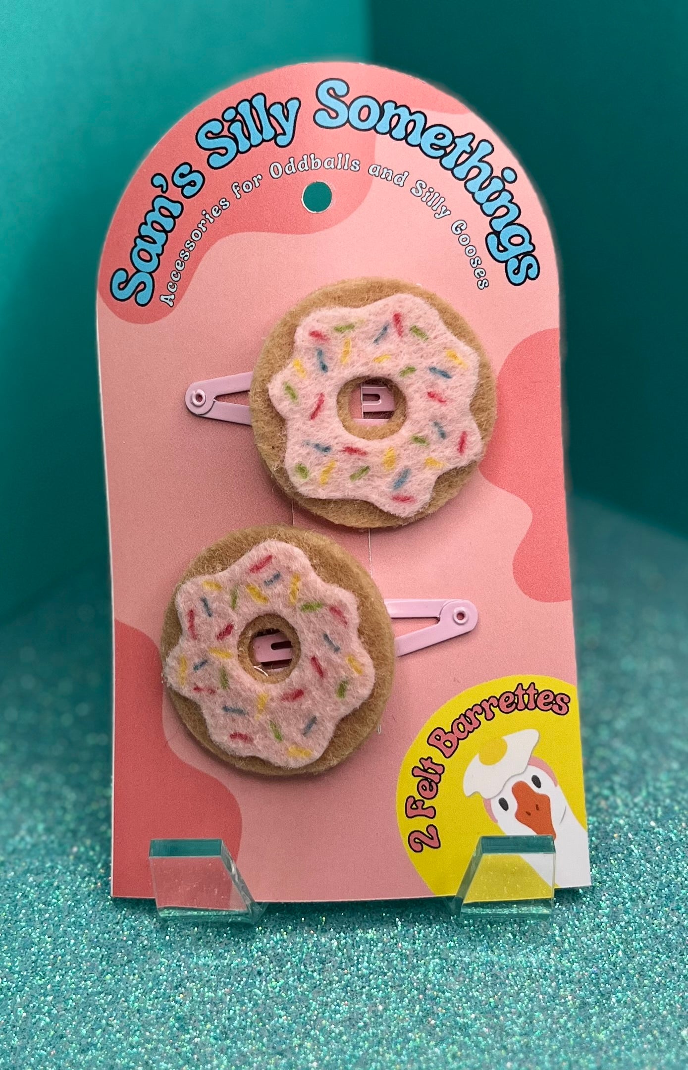 Two donut-shaped hair barrettes with pink icing and sprinkles, attached to a pink cardboard backing with the text 'Sam's Silly Somethings'.