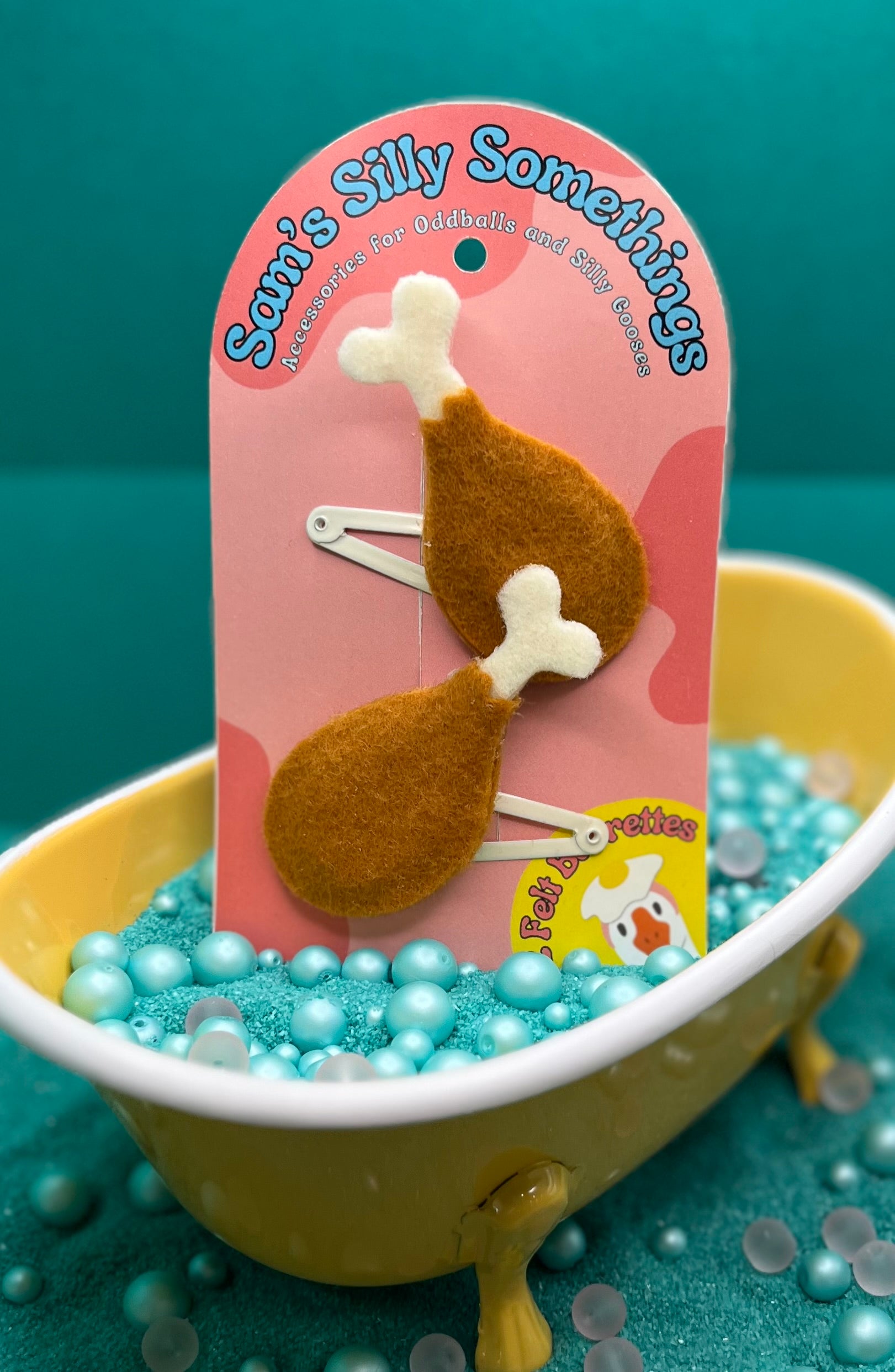 Two brown drumstick-shaped barrettes on a pink packaging card, presented in a yellow bathtub filled with blue beads, simulating water.