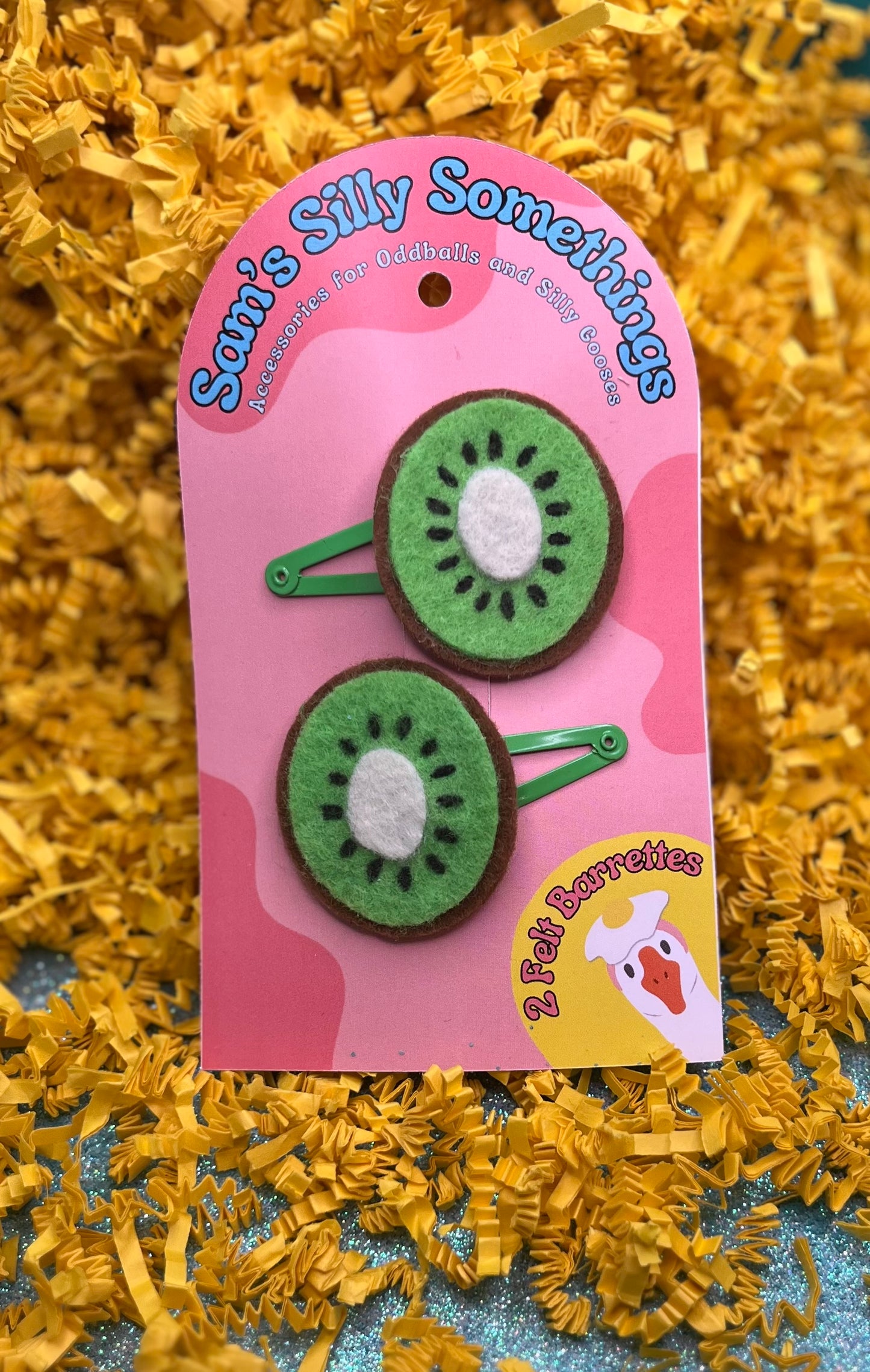 Two green kiwi slice barrettes attached to a pink clip, packaged on a card with yellow decorative elements.