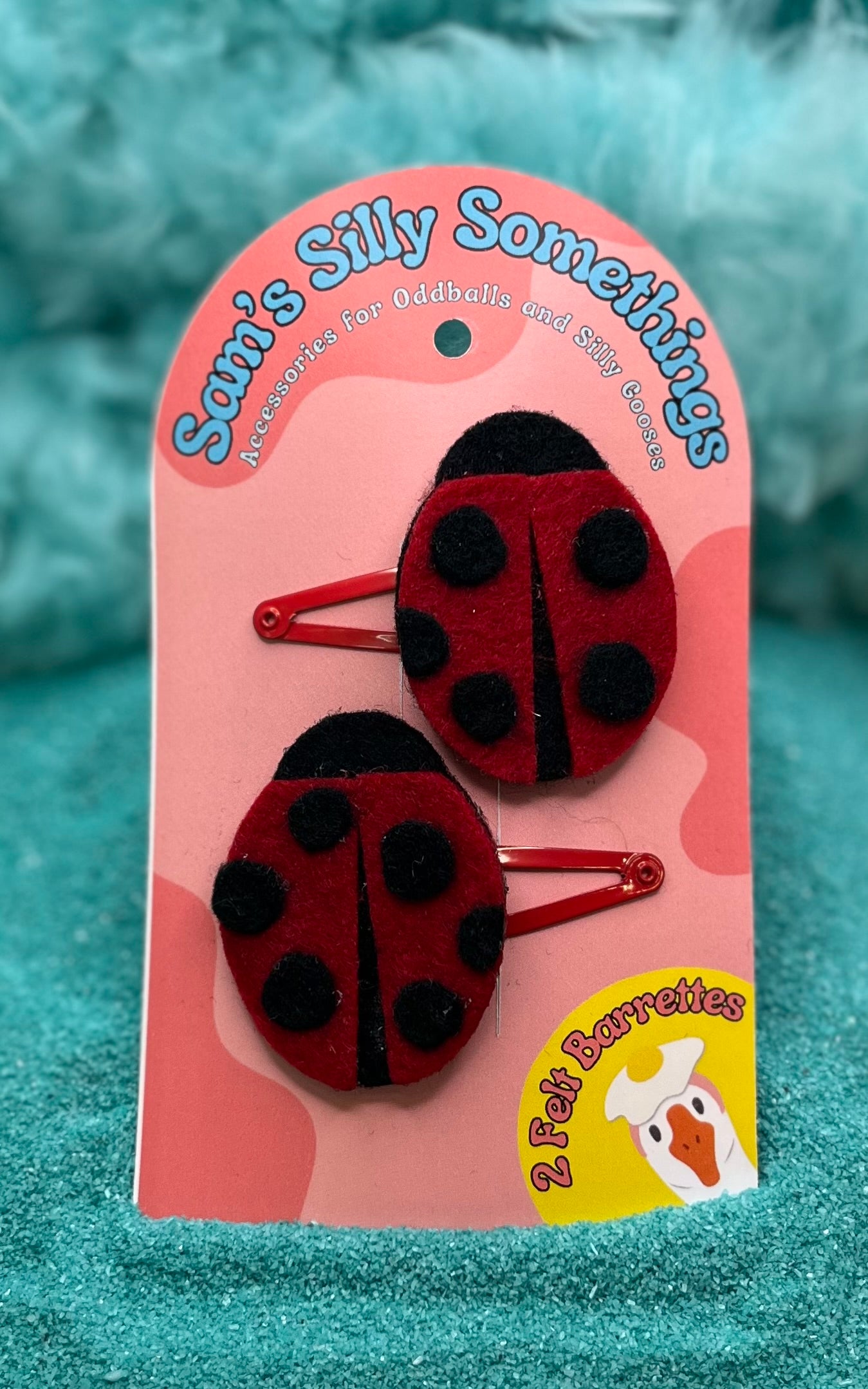 Two red ladybug barrettes with black spots, attached to a pink cardboard backing with a playful design and text that reads 'Sam's Silly Somethings.'.