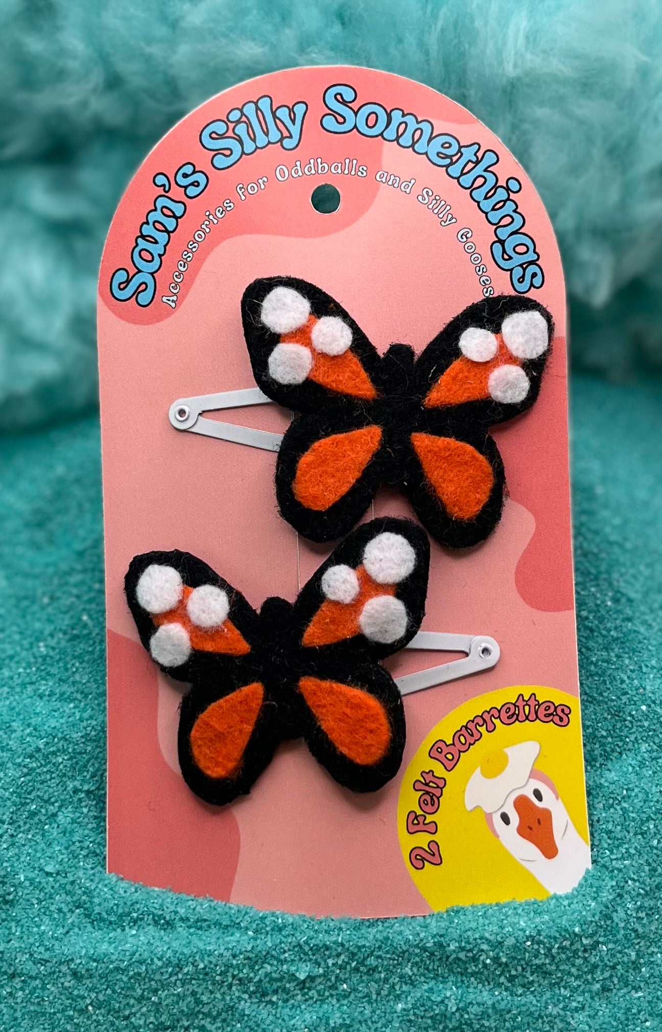Two butterfly-shaped hair barrettes with black, orange, and white colors, attached to a pink cardboard backing with the text 'Silly's Silly Somethings'.