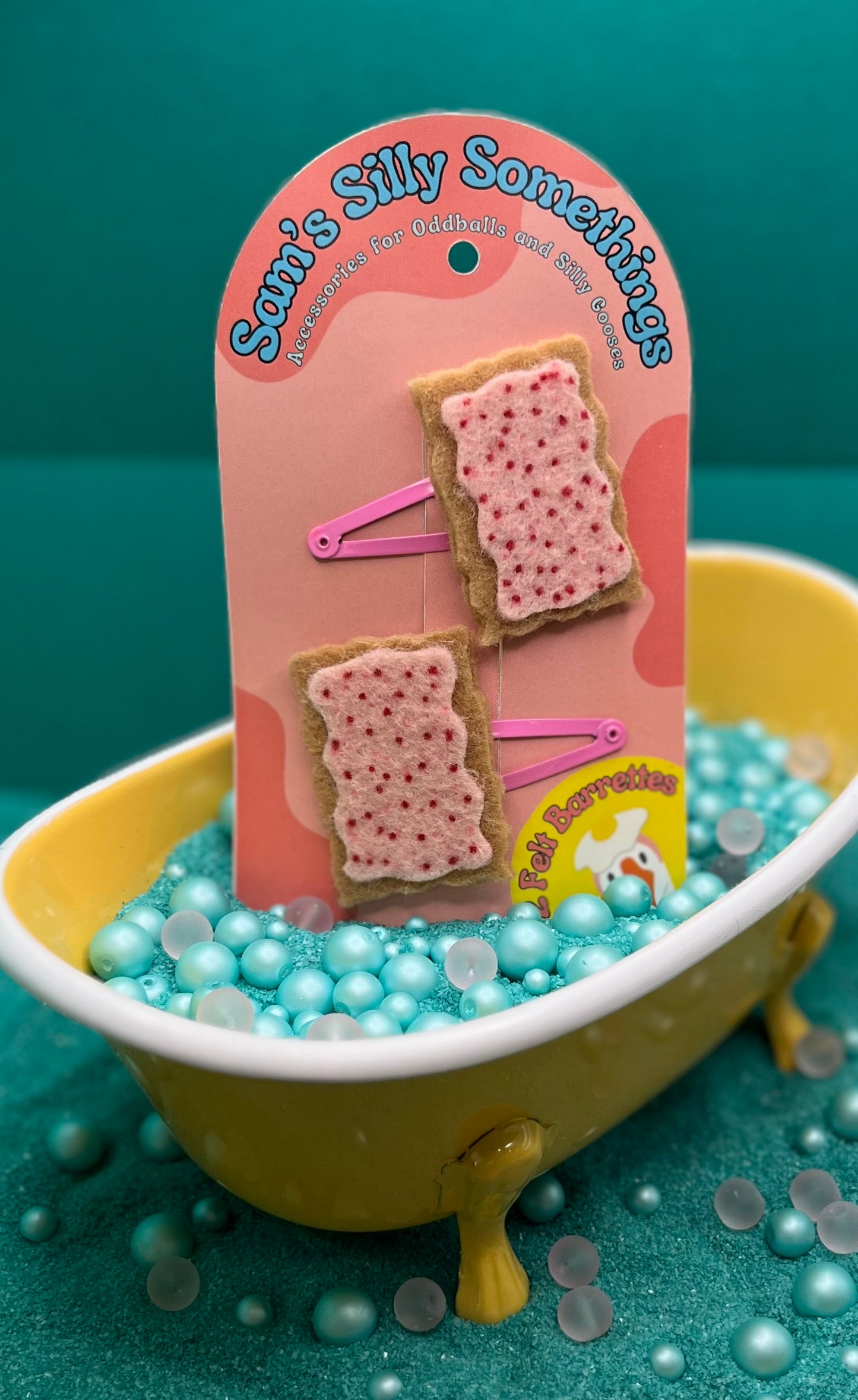 Two barrettes designed to look like pop-tarts placed in a bathtub filled with toy beads, against a pink background with packaging that reads 'Sam's Silly Something'.