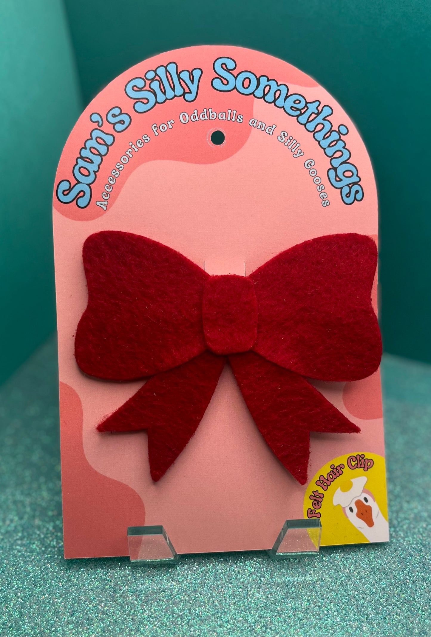 Red bow hair clip on a pink 'Sam's Silly Somethings' branded card against a teal background