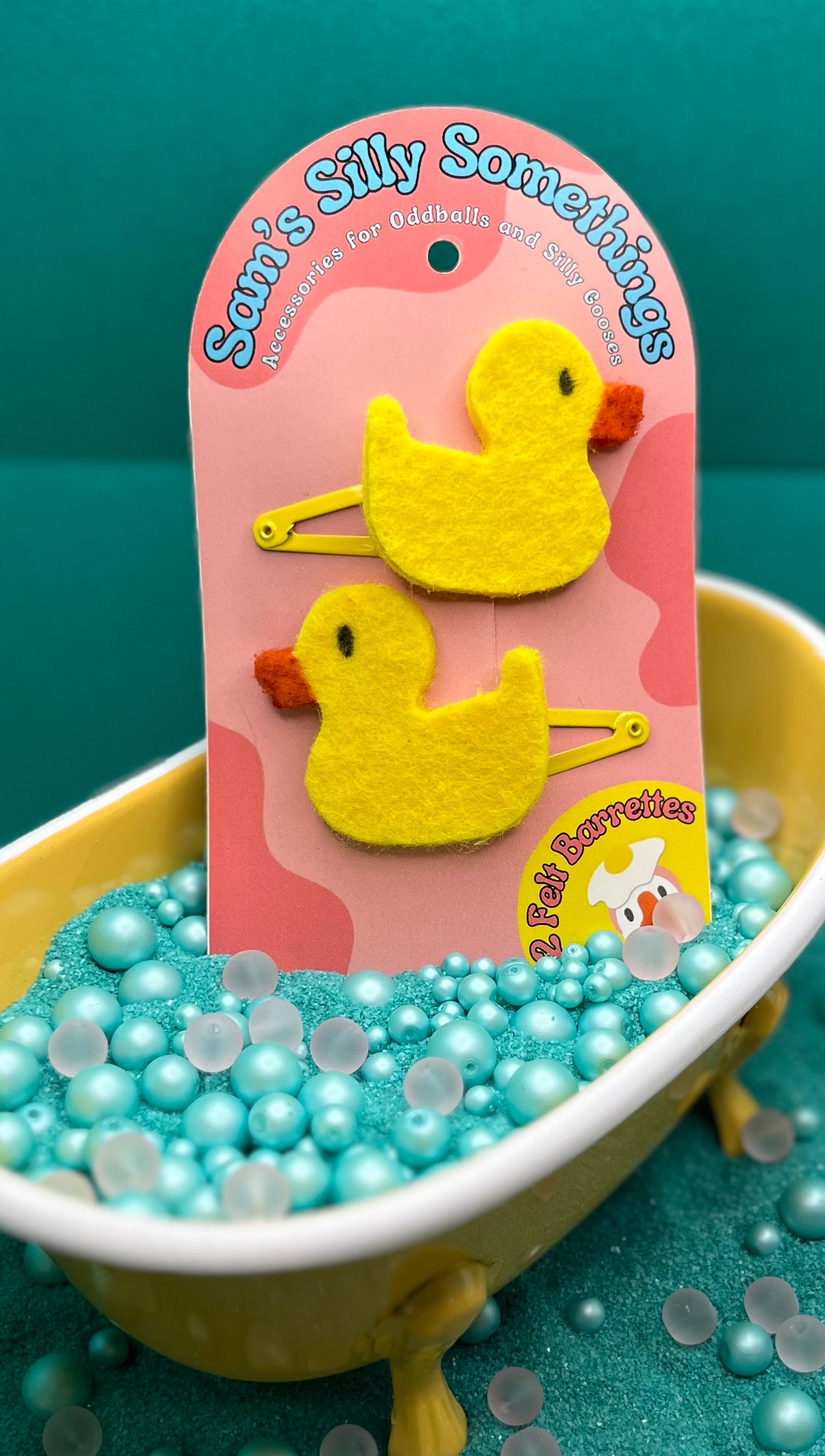 Yellow duck-shaped hair clips on a pink card with 'Sam's Silly Something' branding, placed in a small bathtub filled with blue beads.