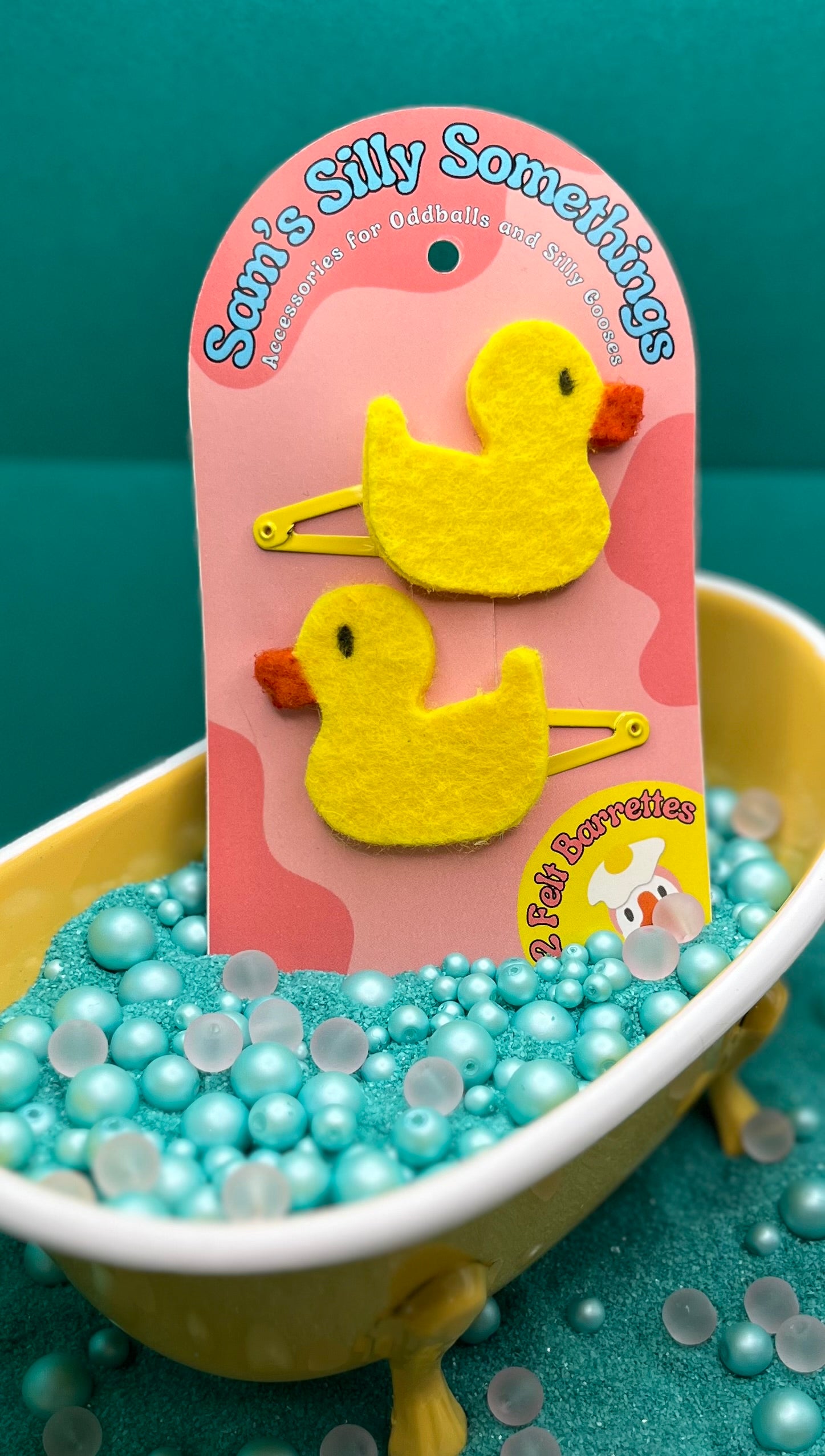 Yellow duck-shaped hair clips on a pink card with 'Sam's Silly Something' branding, placed in a small bathtub filled with blue beads.
